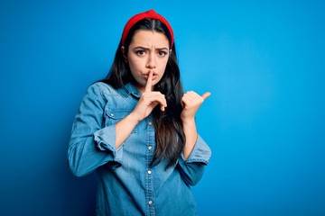 Fototapeta premium Young brunette woman wearing casual denim shirt over blue isolated background asking to be quiet with finger on lips pointing with hand to the side. Silence and secret concept.