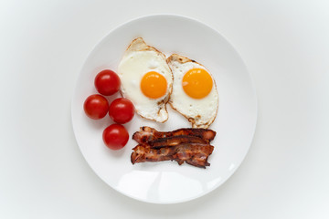 White plate with two fried eggs, crispy bacon and fresh cherry tomatoes on the white table. Top view.