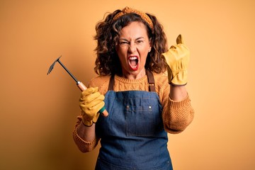 Middle age gardener woman wearing apron holding rake over isolated yellow background annoyed and...
