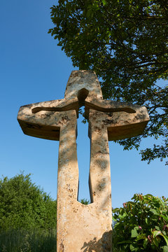 Large and brown cross made of stone, carved religious figure, green nature and blue sky in the background, Germany.