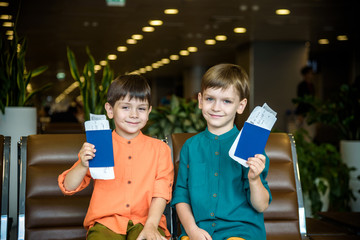 Two young kids sitting on brown chairs, holding passports and tickets in waiting hall in airport. Travel and holidays with children concept