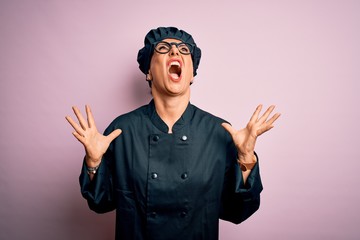 Middle age brunette chef woman wearing cooker uniform and hat over isolated pink background crazy and mad shouting and yelling with aggressive expression and arms raised. Frustration concept.