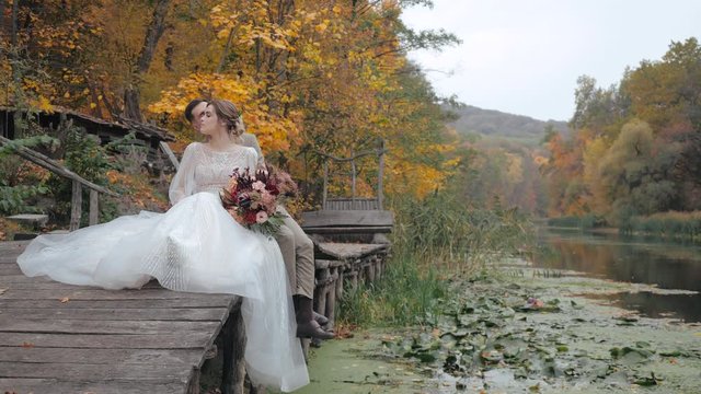 Frontal medium shot of young european bride in luxurious wedding dress and with bouquet of flowers sitting on wooden bridge and hugging and kissing from behind her bridegroom by the river in the fall.