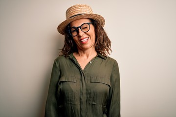 Middle age brunette woman wearing glasses and hat standing over isolated white background winking looking at the camera with sexy expression, cheerful and happy face.