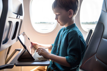 Cute six years old boy, playing on tablen in aircraft on boar, traveling on vacation with parents and siblings