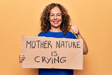 Fototapeta na wymiar Middle age woman asking for environment holding banner with mother nature is crying message smiling happy and positive, thumb up doing excellent and approval sign