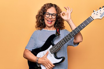 Middle age beautiful musician woman playing electric guitar over isolated yellow background doing...
