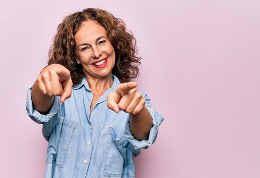 Middle age beautiful woman wearing casual denim shirt standing over pink background pointing to you and the camera with fingers, smiling positive and cheerful