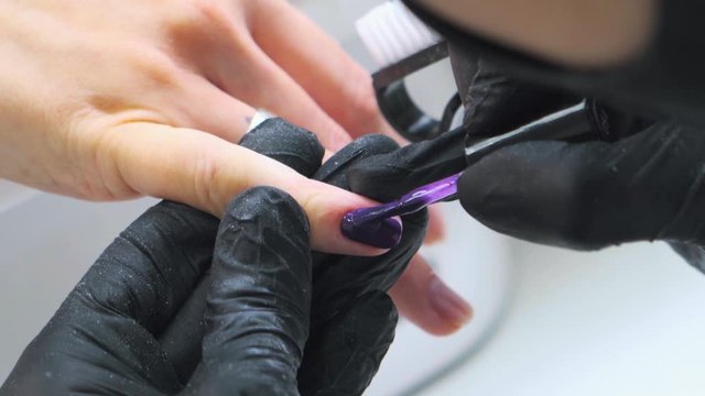 Nails artist is applying purple nail polish during manicure. Nail artist is wearing black gloves and black face mask.