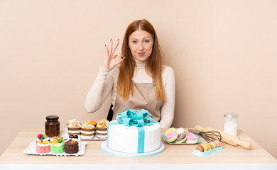 Obraz na płótnie Canvas Young redhead woman with a big cake showing an ok sign with fingers