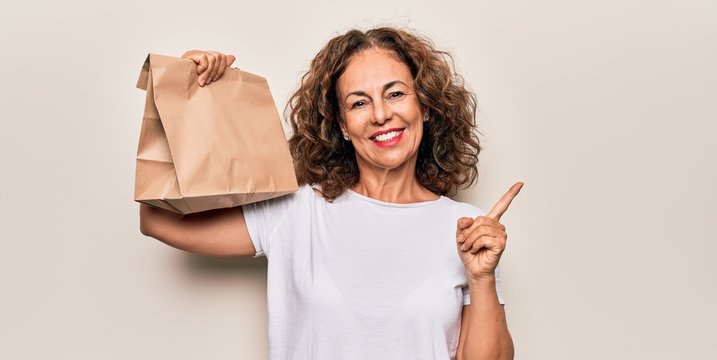 Middle age woman holding deliver paper bag with takeaway food over white background smiling happy pointing with hand and finger to the side