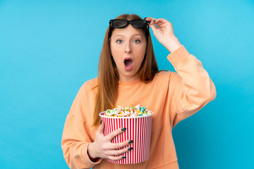 Young redhead woman over isolated background surprised with 3d glasses and holding a big bucket of popcorns