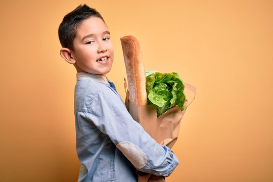 Adorable toddler holding paper bag with food standing over isolated yellow background