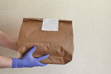 Male hands in blue latex glove gives paper bag to female hands. Closeup of male hands in gloves hold brown craft paper bag with blank white sticker. Delivery service concept. Copy space area.