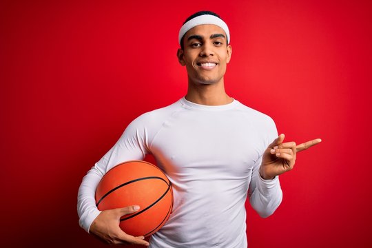 Young handsome african american sportsman holding basketball ball over red background very happy pointing with hand and finger to the side