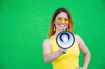 Fototapeta premium Happy woman in a yellow dress glasses and earrings on a green background shouting into a megaphone. Portrait of a girl holding a loudspeaker. Lady with a perfect snow-white smile.