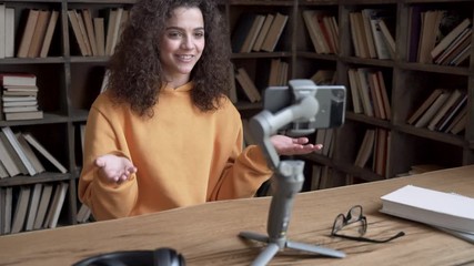 Smiling hispanic gen z teen girl social media channel blogger content creator recording vlog video tutorial on smartphone cam in library. School student vlogger talk look at mobile phone shoot blog.