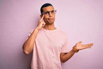 Handsome african american man wearing casual t-shirt and glasses over pink background confused and annoyed with open palm showing copy space and pointing finger to forehead. Think about it.