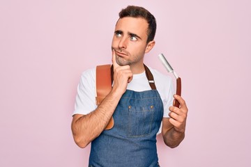 Handsome barber man with blue eyes wearing apron using razor blade over pink background serious...