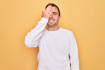 Young handsome man with blue eyes wearing casual sweater standing over yellow background covering one eye with hand, confident smile on face and surprise emotion.