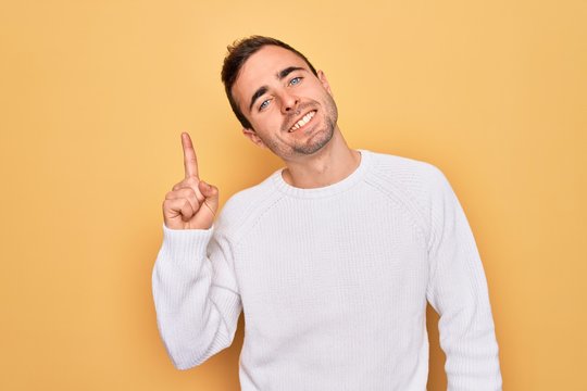 Young handsome man with blue eyes wearing casual sweater standing over yellow background pointing finger up with successful idea. Exited and happy. Number one.