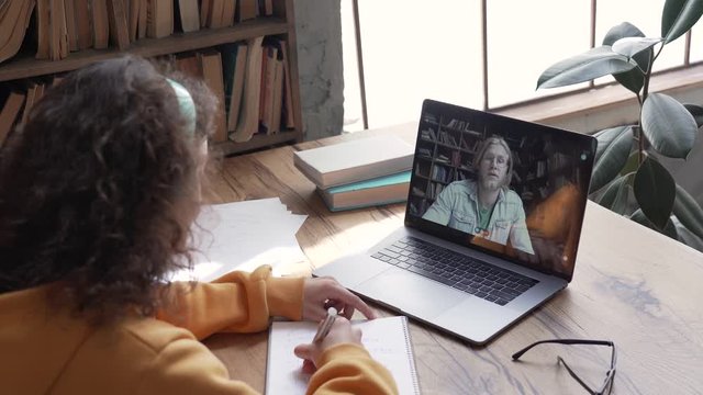 Hispanic teen girl school college student study distance learning talk with online teacher on laptop screen write notes. Elearning zoom video call, videoconference class with tutor. Over shoulder view