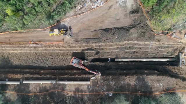 Aerial drone above construction site outside at nature with green trees around. Contractor digging with crane and shovel a water channel placing and installing cement pipelines tubes
