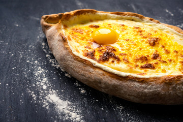 Khachapuri adjara traditional Georgian cuisine meal. Baked bread as boat with cheese and egg filling on black background, decorated with flour. National pastry snack.