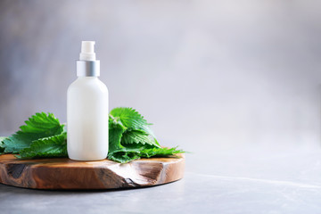 Obraz na płótnie Canvas Nettle lotion, cream, shampoo or soap in white bottle and fresh nettles leaves on grey background. Medicinal herb for health and beauty, skin care and hair treatment.