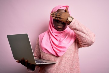 African american plus size woman wearing muslim hijab using laptop over pink background covering eyes with hand, looking serious and sad. Sightless, hiding and rejection concept