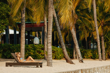 Beautiful young lady in blue trendy swimsuit lying on sunbed under palm trees. Happy tourist in...