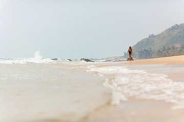 Back view of young woman in black bikini walking in distance among seashore. Beautiful girl enjoying summer vacation on Hainan island. Tropical nature around.
