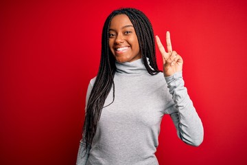 Young african american woman wearing turtleneck sweater over red isolated background smiling with happy face winking at the camera doing victory sign. Number two.