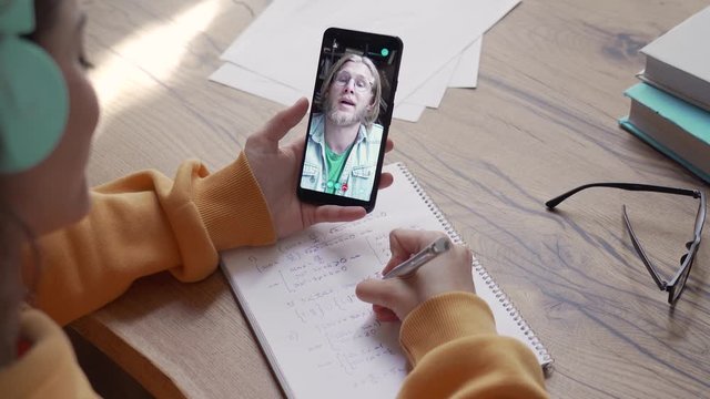 Teen girl school college student wear headphones hold phone distance learn online class with math teacher tutor using mobile zoom app video conference call app write notes. Over shoulder closeup view.