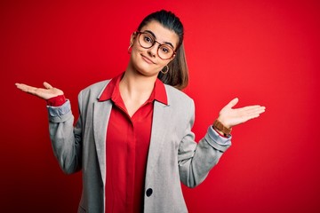 Young beautiful brunette businesswoman wearing jacket and glasses over red background clueless and...