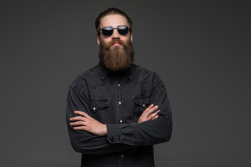 Portrait of handsome bearded young man with serious expression wearing sunglasses with crossed hands over gray background