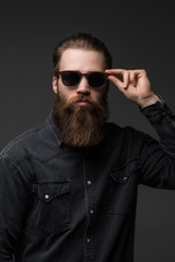 Obraz premium Portrait of handsome s bearded young man with serious expression wearing sunglasses over gray background