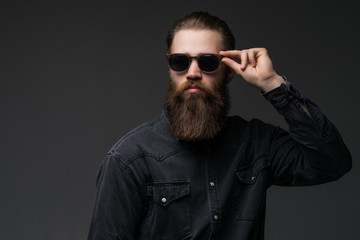 Portrait of handsome s bearded young man with serious expression wearing sunglasses over gray...