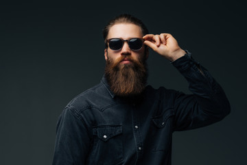 Obraz premium Portrait of handsome s bearded young man with serious expression wearing sunglasses over gray background