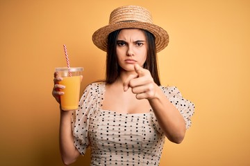 Young beautiful brunette woman on vacation wearing summer hat drinking orange juice pointing with finger to the camera and to you, hand sign, positive and confident gesture from the front