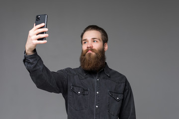 Happy smiling hipster man with long beard taking selfie with hands on camera standing on gray background