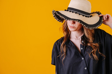 Close up portrait of a young woman holding straw hat low, so it's covering her eyes. She has long brown hair. Over orange background.