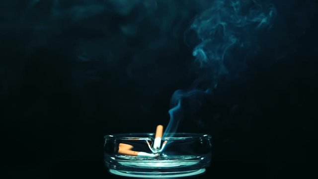 A cigarette lies in an ashtray and smokes, smoke disperses around, next to it lies a already smoked cigarette, on a black background. Closeup. The camera moves around. Slow motion. High speed camera