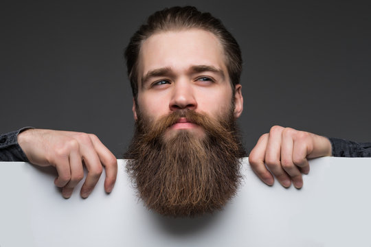 Young long bearded man over blank text on white sheet on gray background.