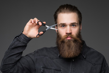 Young bearded man holding barber scissors and razor near his face standing isolated on gray background