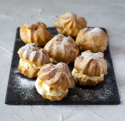 Cream puffs or profiteroles dusted with sugar powder and and filled with whipped cream on black slate board