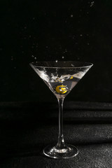 Martini cocktail with olive splash and drops on black background with shadows on table. Copy space. Alcohol drink in a glass.