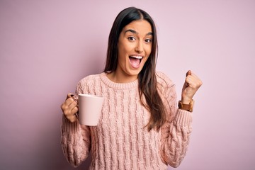 Young beautiful brunette woman drinking cup of coffee over isolated pink background screaming proud and celebrating victory and success very excited, cheering emotion