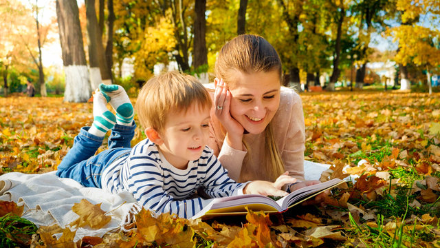 Portrait of happy smiling boy with mother reading book on picnic at autumn park
