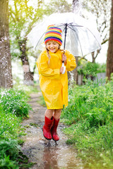 little Blonde Girl Smiles Jumps on Puddles in Spring in yellow raincoat and rubber boots with...